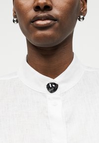 Γυναίκα που φοράει λευκό πουκάμισο με γιακά, με μαύρο διακοσμητικό κουμπί και στρογγυλά ασημένια σκουλαρίκια, εμφανίζεται από το πηγούνι μέχρι την κλείδα.