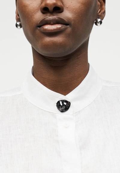 Femme portant une chemise blanche à col avec un bouton décoratif noir et des boucles d'oreilles rondes en argent, montrée du menton à la clavicule.
