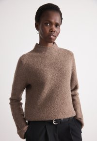 Pullover castanho texturizado com gola alta e mangas longas, combinado com calças pretas e um cinto com uma fivela prateada.