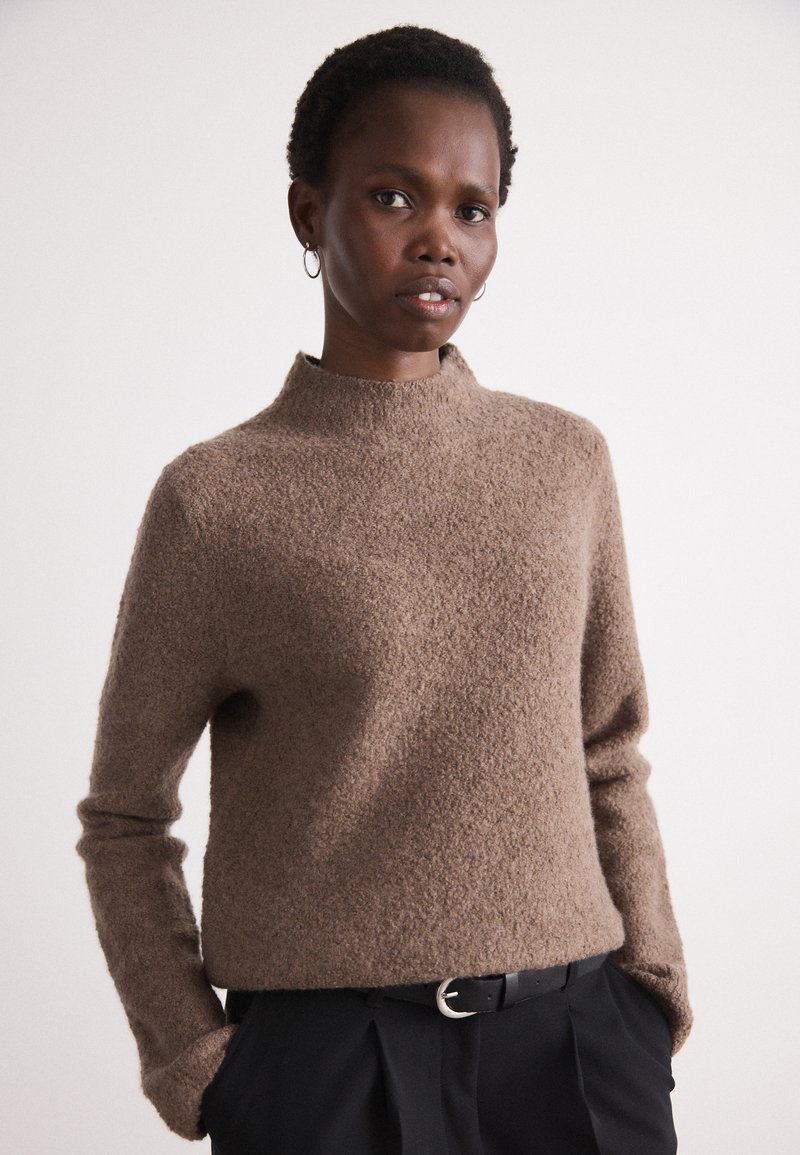 Pullover castanho texturizado com gola alta e mangas longas, combinado com calças pretas e um cinto com uma fivela prateada.