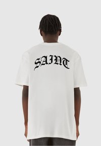 Hvid t-shirt med stort sort tekst der står "SAINT" på ryggen. Korte ærmer, rund halsudskæring og en afslappet pasform.