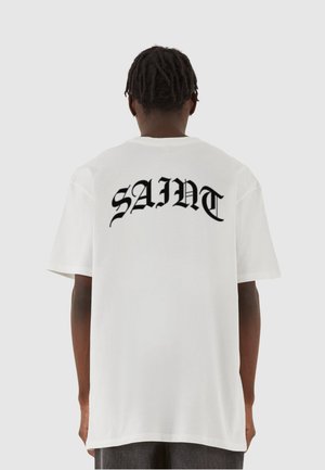 Hvid t-shirt med stort sort tekst der står "SAINT" på ryggen. Korte ærmer, rund halsudskæring og en afslappet pasform.