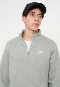 Šedá mikina na zip vyrobená z měkké látky, s vysokým límcem a malým bílým logem Nike na hrudi. Nosená s černou čepicí.