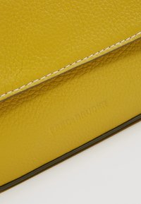 Žlutá kožená taška s texturovaným povrchem. Má bílé švy a vystouplý detail loga. Obdélníkový tvar s elegantním designem.