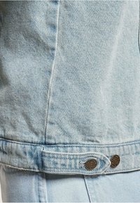 Světle modrá džínsová bunda s texturovaným povrchem, dvěma kovovými knoflíky na lemu a vyšívanými detaily podél švů.