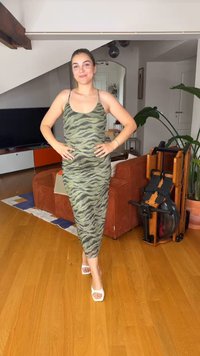 Νεαρή γυναίκα με πράσινο φόρεμα με σχέδιο ζέβρας στέκεται με τα χέρια στη μέση σε ένα σαλόνι με ξύλινο πάτωμα και πορτοκαλί καναπέ.