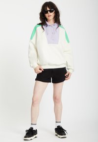 Sweatshirt mit großem lila Vorderteil und grünen Schulterakzenten, aus weichem Material gefertigt; kombiniert mit schwarzen Shorts und Turnschuhen.