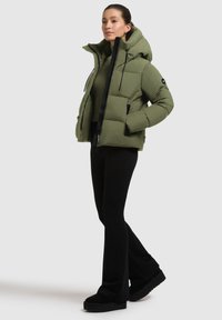 Donna con giacca trapuntata verde oliva con cappuccio, pantaloni neri a campana e scarpe con tacco nere, in piedi contro uno sfondo bianco.