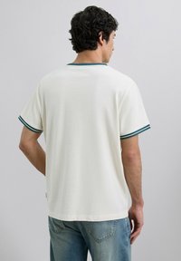 Camiseta blanca de manga corta con rayas verdes en los puños y el cuello. Textura suave con un ajuste relajado, combinada con jeans azules descoloridos.