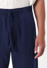 Pantalons bleu marine texturés avec une taille élastique et un cordon de serrage ajustable, présentant une coupe décontractée. Placement des mains visible près du côté.