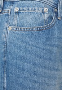 Jeans de mezclilla azul claro con una vista en primer plano de un bolsillo trasero, que presenta herrajes plateados, costuras en contraste y un acabado ligeramente texturizado.