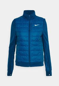 Veste matelassée bleue avec col haut, fermeture éclair frontale, poignets élastiques et logo Nike sur le côté gauche. Caractérisée par une texture matelassée.