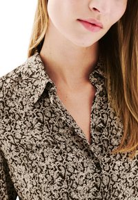 Blouse marron et crème à motifs avec un col, comportant des détails de boutons et une texture douce, partiellement visible sur le haut du corps d'un mannequin.