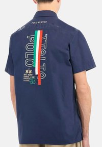 Marineblauw shirt met korte mouwen met de tekst "POLO ITALIA", groen, wit en rode strepen, en geborduurd paardenrijder op de achterkant, gedragen door een persoon.