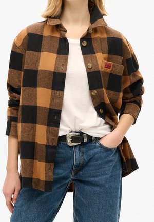 Person trägt eine braun-schwarz karierte Jacke über einem weißen T-Shirt, blaue Jeans mit schwarzem Gürtel, eine Hand in der Tasche.