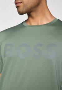 Homme portant un t-shirt à col rond vert avec le logo "BOSS" sur la poitrine, montrant un profil latéral partiel et le cou.