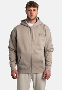 Sudadera con cremallera de color beige, con una gran capucha, bolsillos frontales y puños acanalados. Presenta un logotipo circular en el lado izquierdo del pecho. Tejido de textura suave.
