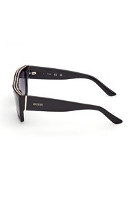 Lunettes de soleil Guess noires avec montures rectangulaires épaisses et verres foncés, ornées d'un logo doré et de strass sur le bord supérieur.