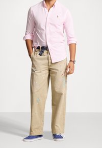 Ljust rosa knappad skjorta, beige målarsbyxor med färgstänk och blå canvas-skor; med ett randigt bälte som accent.