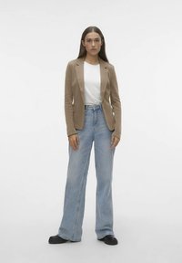 Beige getailleerde blazer over een wit T-shirt, gecombineerd met lichtblauwe wijde broekspijpen en zwarte schoenen, staand tegen een effen achtergrond.