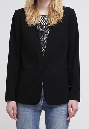 Kvinde iført en sort blazer over en sort blondeoverdel og lyseblå jeans, stående mod en ensfarvet lysegrå baggrund.