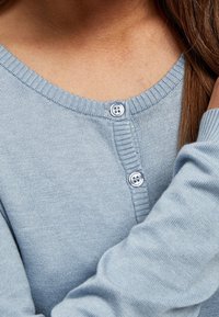 Ljusblå cardigan med ribbad halsringning, med två runda, ljusa knappar framtill. Mjuk textur och åtsittande passform.