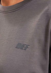 Gray sweatshirt featuring a textured fabric, round neckline, and a black "DEF" logo on the left side. Clean design with minimal detailing.