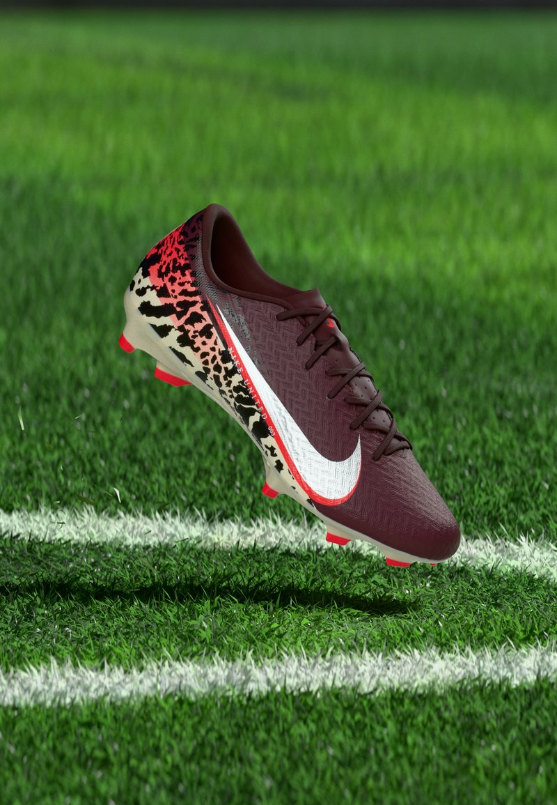 Μπότες ποδοσφαίρου σε μαροόν και εκρού χρώμα με κόκκινο και μαύρο πουά σχέδιο, λογότυπο swoosh σε λευκό, αιωρούμενες πάνω από πράσινο χορτάρι κοντά σε λευκή γραμμή γηπέδου.
