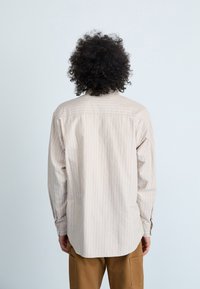 Beige stribet langærmet skjorte lavet af let materiale, med en buet kant og knapmanchetter, vist fra baggrunden.