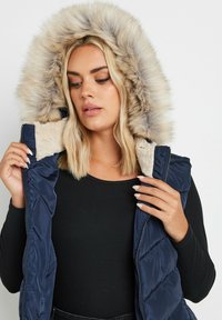 Laivastonsininen puffatakkivesti, jossa on tekoturkishuppu, vetoketjukiinnitys ja pehmeä sisävuori. Varustettu tikatulla pinnalla ja sivutaskuilla.