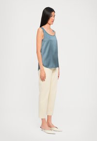 Woman standing in cream trousers, blue sleeveless satin top, and cream low-heeled pointed shoes against a plain white background.