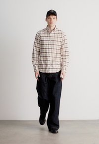 Camicia a quadri beige con linee marroni e viola, abbinata a pantaloni di jeans neri larghi e scarpe nere. Un cappello nero completa il look.