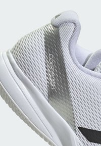 Chaussure de sport blanche avec un dessus en mesh texturé, des accents argentés et une semelle en caoutchouc. Présente la marque "COURTFLASH" sur le côté.