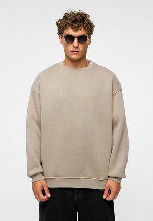 Beige oversized sweatshirt met verlaagde schouders, ronde hals en ribbelboorden. Draagt het met ronde, donkere zonnebrillen en zwarte broek.