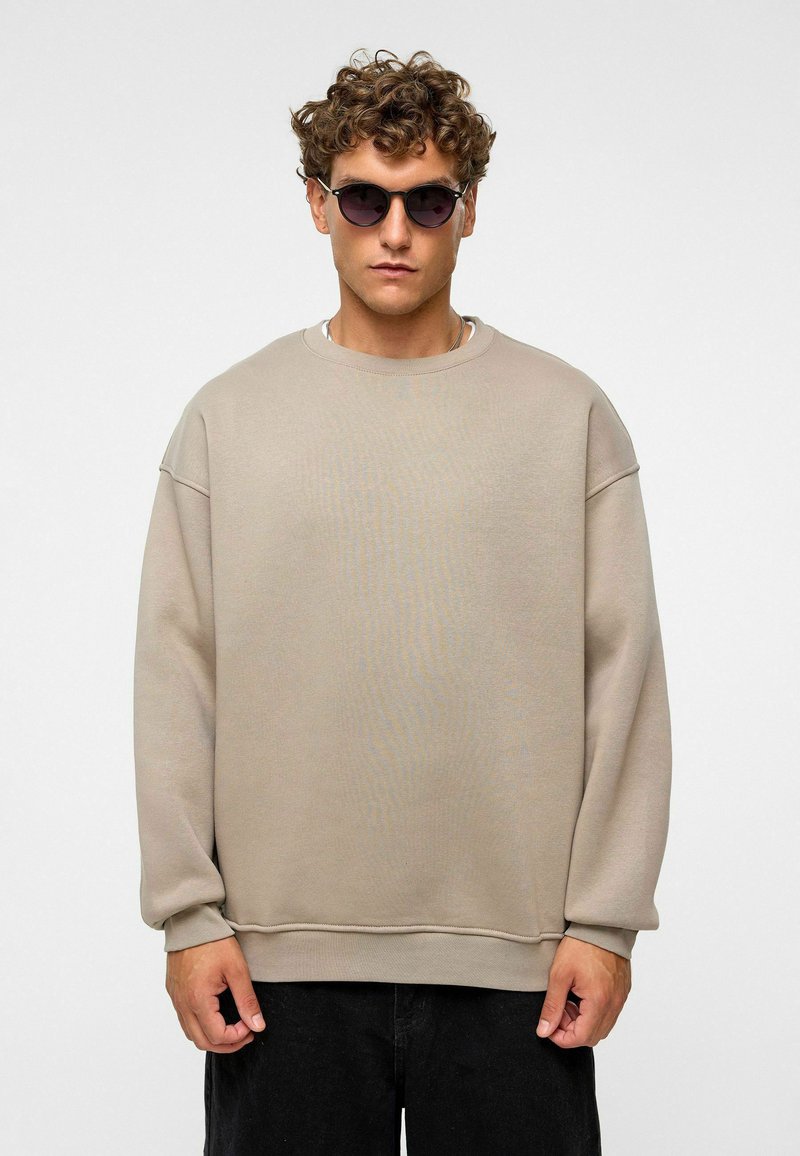 Beige oversized sweatshirt met verlaagde schouders, ronde hals en ribbelboorden. Draagt het met ronde, donkere zonnebrillen en zwarte broek.