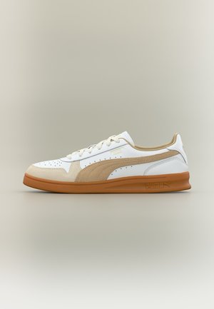 Λευκό και μπεζ παπούτσι Puma χαμηλού προφίλ με οπές, σουέντ λεπτομέρειες και λαστιχένια σόλα, εμφανιζόμενο σε πλευρική όψη.