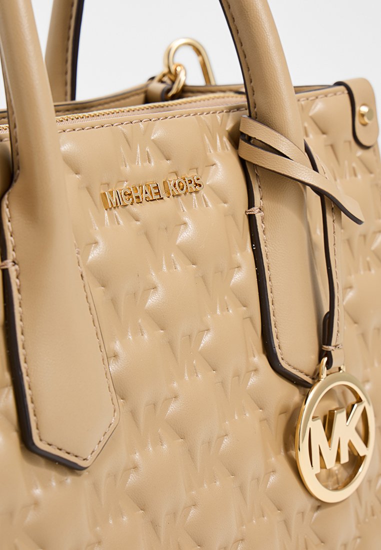 Sac à main en cuir beige avec un motif matelassé, quincaillerie dorée et un charme logo circulaire. Dispose de deux poignées et d'une fermeture supérieure sécurisée.