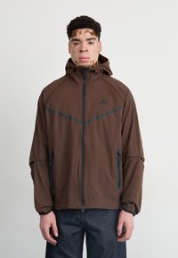 Brown hooded jacket with black accents, zippered pockets, and a lightweight fabric. It features a sleek design and elastic cuffs.