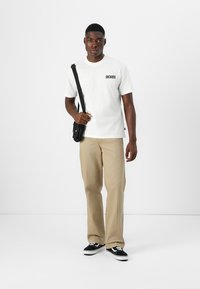 Camiseta blanca con logo negro de "DICKIES", pantalones beige, bolso cruzado negro y zapatillas en blanco y negro, de pie sobre un fondo sencillo.