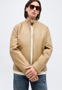 Άνδρας φοράει δερμάτινο τζάκετ ταμπά με φερμουάρ, λευκό πουκάμισο, μπλε τζιν και μεγάλα τετράγωνα γυαλιά ηλίου, στέκεται με τα χέρια ενωμένα.