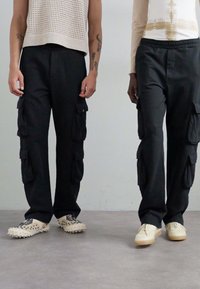 Pantaloni cargo neri dotati di molteplici tasche, vestibilità comoda ed elastico in vita, abbinati a calzature di colore chiaro. Top in maglia con texture visibile.