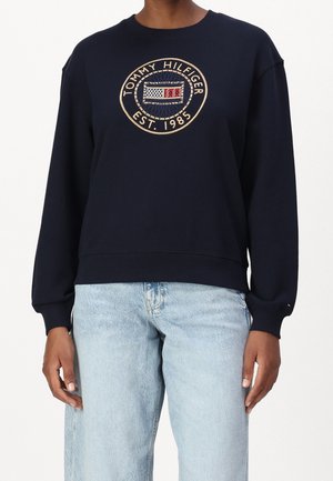 Person iført en navyblå Tommy Hilfiger sweatshirt med logo og tekst "Est. 1985", kombineret med lyseblå jeans.