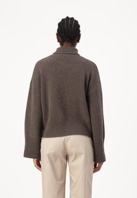 Καφέ πλεκτό πουλόβερ με ψηλό λαιμό, κατεβασμένους ώμους και ριμπ τελείωμα. Φοριέται με ανοιχτόχρωμα παντελόνια, προσφέροντας μια άνετη εφαρμογή και υφή.