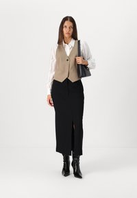 Beige vest over en hvid skjorte med knapper, parret med en sort nederdel med en slidse. Sorte ankelstøvler, holder en lille sort taske.
