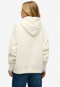 Sweat à capuche crème oversize avec poche kangourou et capuche. Tissu doux et poignets côtelés. Porté par-dessus une chemise rayée bleue et blanche.