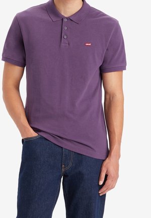 Mann trägt ein kurzärmeliges lila Polohemd mit drei Knöpfen und kleinem rotem Levi’s-Logo, kombiniert mit dunkelblauen Jeans.