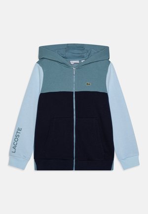 Sweat-shirt à capuche zippé avec manches bleu clair, corps bleu foncé, partie supérieure turquoise, poches avant et logo Lacoste sur la poitrine et la manche.