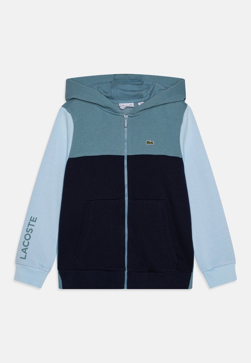 Sweat-shirt à capuche zippé avec manches bleu clair, corps bleu foncé, partie supérieure turquoise, poches avant et logo Lacoste sur la poitrine et la manche.