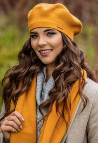 Berete de punto amarillo con una superficie texturizada; bufanda de punto gris enrollada alrededor del cuello; tonos cálidos y terrosos; cabello liso enmarcando el rostro.