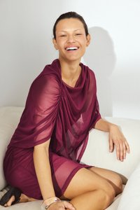 Bordeaux jurk met doorschijnende, gedrapeerde stof, voorzien van geplooide details op de schouders, gedragen met zwarte sandalen en een transparante armband.